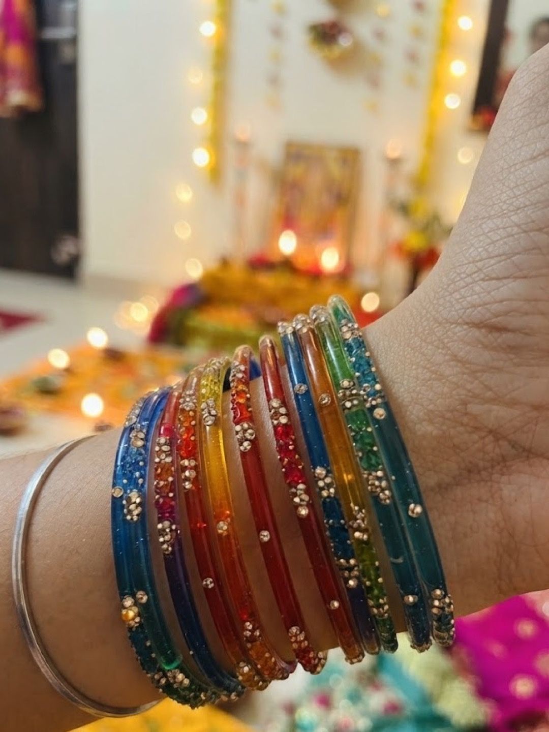 Twelve rainbow chudiyan worn on a wrist, with diyas and festive lights behind